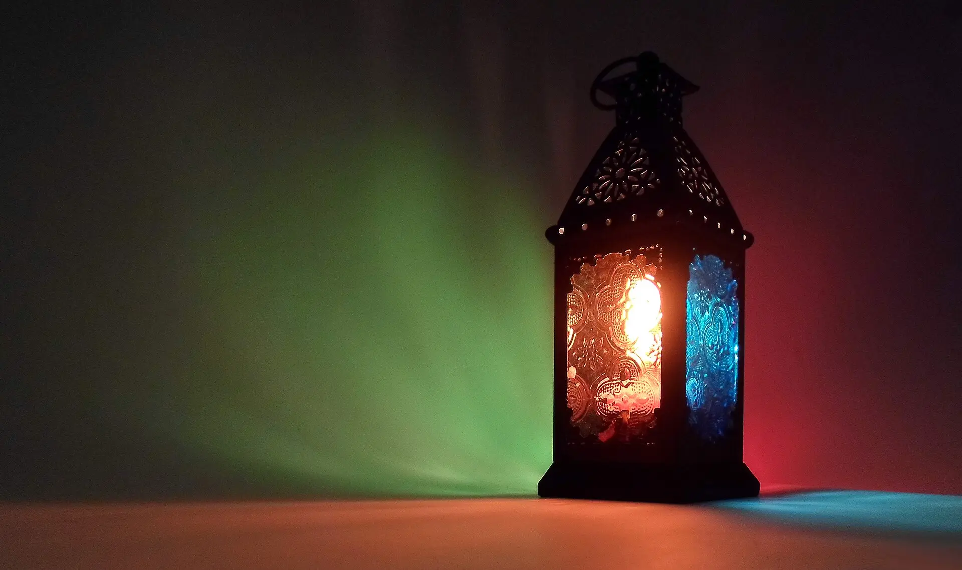 A metal Ramadan lantern with coloured glass panels glowing in a dark room, casting green, red and blue light